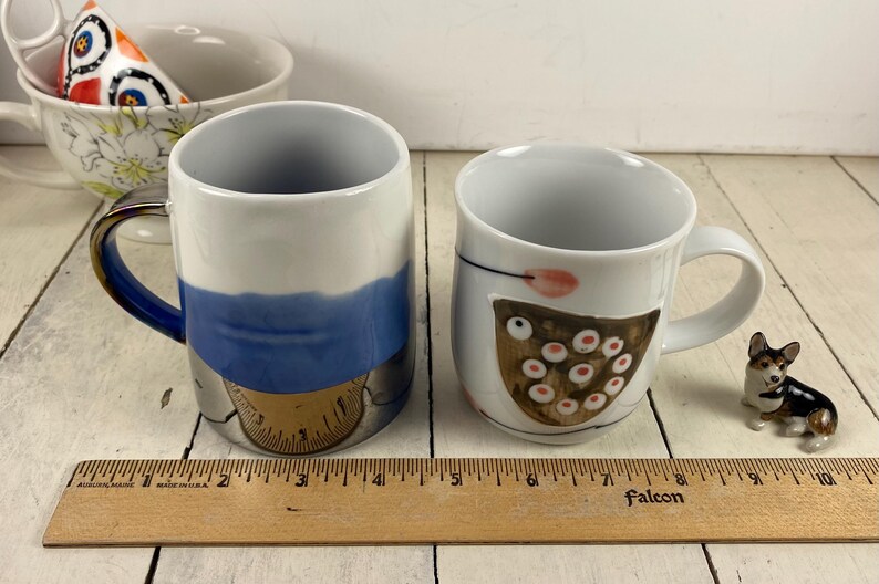K&ouml;nnte beinhalten: Zwei Keramikbecher mit einzigartigen Designs. Ein Becher hat ein blau-wei&szlig;es Design mit goldfarbenem Boden. Der andere Becher ist wei&szlig; mit einem braunen und rosa Design. Eine kleine Hundefigur ist ebenfalls vorhanden.