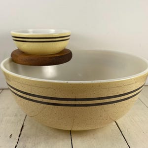 May include: A set of two vintage mixing bowls. The larger bowl is beige with two dark brown stripes and a speckled finish. A smaller bowl, also beige with brown stripes, sits on a wooden coaster. The interior of both bowls is white.