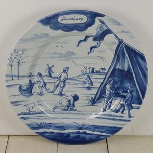 May include: Blue and white ceramic plate with a winter scene depicting people ice skating and a tent. The plate has the word "Januarij" written on it.