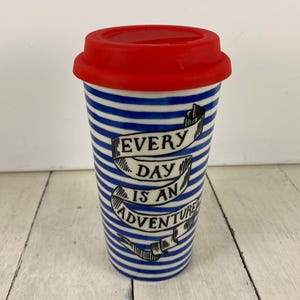 May include: A travel mug with a red lid and a blue and white striped pattern. The mug has the text "EVERY DAY IS AN ADVENTURE" in a banner design. The mug is made of ceramic or a similar material.