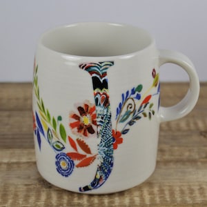 May include: A white ceramic mug with a floral design and the letter "J". The letter is decorated with a colorful pattern. The mug has a curved handle and is set against a wooden surface.