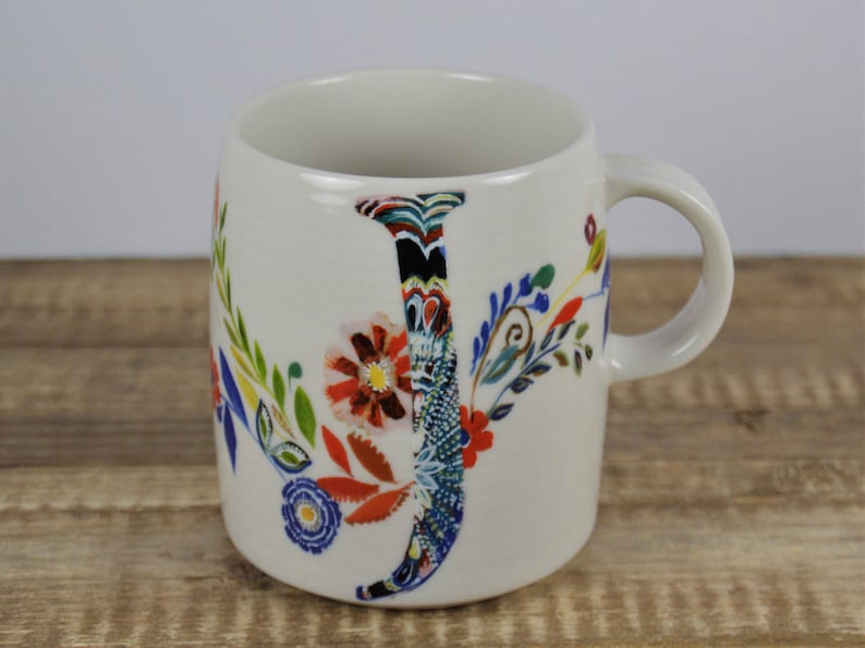 May include: A white ceramic mug with a floral design and the letter "J". The letter is decorated with a colorful pattern. The mug has a curved handle and is set against a wooden surface.