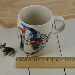 K&ouml;nnte beinhalten: Eine wei&szlig;e Keramiktasse mit einem floralen Design, das den Buchstaben "D" in Blau, Rot, Gelb und Gr&uuml;n zeigt. Die Tasse steht auf einer wei&szlig;en Holzoberfl&auml;che neben einer kleinen Keramik-Hundefigur.