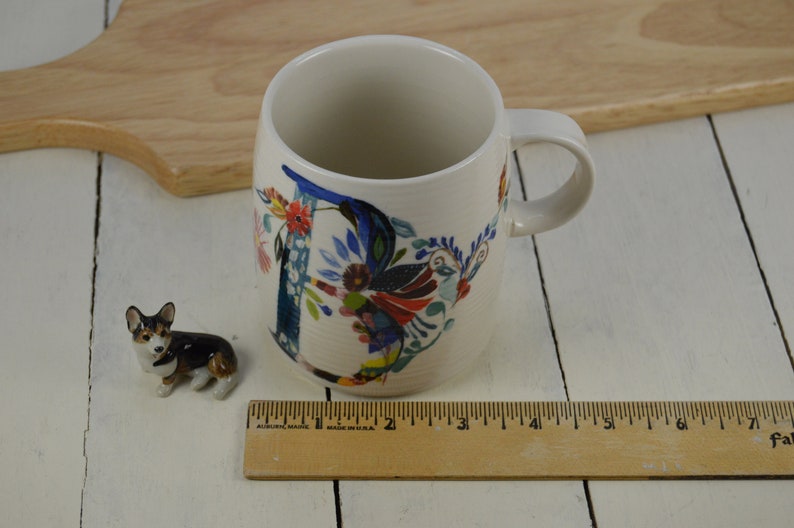 K&ouml;nnte beinhalten: Eine wei&szlig;e Keramiktasse mit einem floralen Design, das den Buchstaben "D" in Blau, Rot, Gelb und Gr&uuml;n zeigt. Die Tasse steht auf einer wei&szlig;en Holzoberfl&auml;che neben einer kleinen Keramik-Hundefigur.