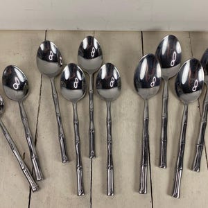 May include: A collection of stainless steel spoons with bamboo-style handles. The spoons are arranged on a light-colored wooden surface. The spoons have a shiny, reflective surface.
