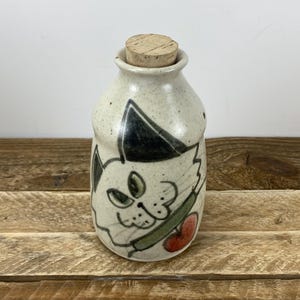 May include: A speckled ceramic bottle with a cork stopper, featuring a hand-painted cat design. The cat has black ears, green eyes, and a red heart. The bottle is a cream color.