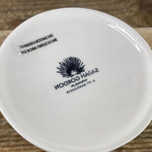 K&ouml;nnte beinhalten: Ein wei&szlig;er Keramikbecherboden mit blau-wei&szlig;em Logo und Text. Der Text lautet "SARAH GORDON" und "DISHWASHER & MICROWAVE SAFE MADE IN CHINA / FABRIQU&Eacute; EN CHINE". Der Becher steht auf einer Holzoberfl&auml;che.