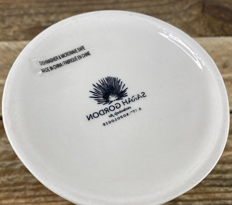 K&ouml;nnte beinhalten: Ein wei&szlig;er Keramikbecherboden mit blau-wei&szlig;em Logo und Text. Der Text lautet "SARAH GORDON" und "DISHWASHER & MICROWAVE SAFE MADE IN CHINA / FABRIQU&Eacute; EN CHINE". Der Becher steht auf einer Holzoberfl&auml;che.