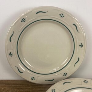 May include: Off-white ceramic plates with a green floral and curved line design. The plates have a green border and a decorative pattern around the rim. The plates are likely for dining and are a classic style.