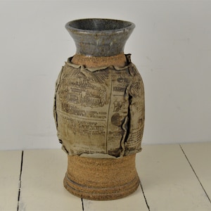 May include: A ceramic vase with a brown and beige glaze. The vase is decorated with a newspaper print design. The vase has a wide mouth and a narrow base.