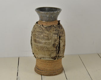 May include: A ceramic vase with a brown and beige glaze. The vase is decorated with a newspaper print design. The vase has a wide mouth and a narrow base.