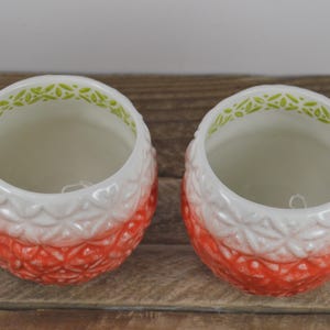 May include: Two red and white ceramic mugs with a green and white floral design around the rim. The mugs are shaped like pineapples.
