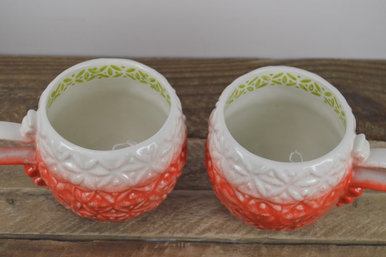 May include: Two red and white ceramic mugs with a green and white floral design around the rim. The mugs are shaped like pineapples.