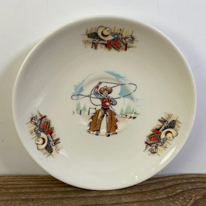 May include: A round white ceramic saucer featuring a central illustration of a cowboy swinging a lasso, surrounded by three smaller decorative motifs of cowboy hats and leather gear arranged symmetrically around the inner rim of the dish.