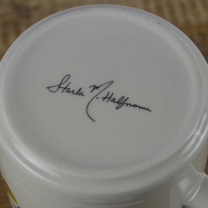May include: White ceramic mug with a curved handle. The bottom of the mug features a handwritten signature in black ink. The mug's interior is white, and the exterior has a subtle, textured design. The mug is set against a wooden surface.