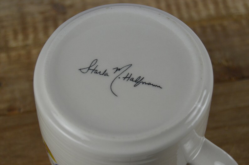 May include: White ceramic mug with a curved handle. The bottom of the mug features a handwritten signature in black ink. The mug's interior is white, and the exterior has a subtle, textured design. The mug is set against a wooden surface.