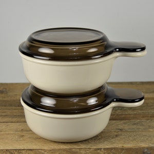 May include: Two white ceramic bowls with brown glass lids and handles. The bowls are stacked on top of each other.