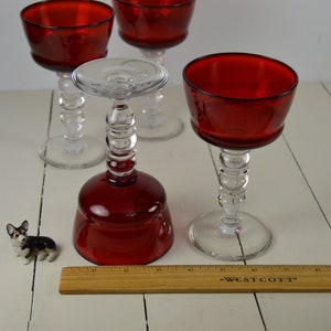 May include: Four red glass wine glasses with clear stems and bases. The glasses have a beaded design on the stems. A small ceramic dog figurine is in the foreground, and a wooden ruler is in the bottom of the image.