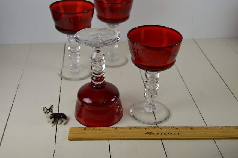 May include: Four red glass wine glasses with clear stems and bases. The glasses have a beaded design on the stems. A small ceramic dog figurine is in the foreground, and a wooden ruler is in the bottom of the image.