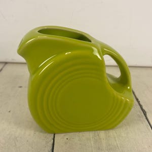 May include: A lime green ceramic pitcher with a rounded body and a handle. The pitcher has a spout for pouring and a circular design on the front. The pitcher is made of ceramic and has a glossy finish.