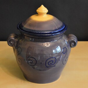 May include: A purple ceramic jar with a yellow lid and blue swirl designs. The jar has two handles and a knob on the lid.