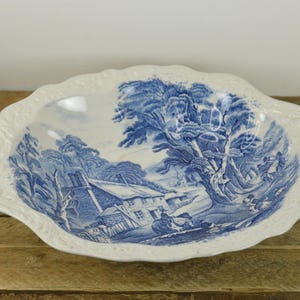 May include: A white ceramic bowl with a blue and white design of a cottage in a forest. The bowl has a scalloped edge and is perfect for serving.