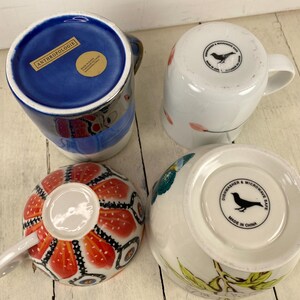 K&ouml;nnte beinhalten: Vier Keramikbecher mit verschiedenen Designs. Ein Becher hat ein blaues Innenleben mit dem Text "ANTHROPOLOGIE". Ein weiterer hat ein rot-oranges Blumenmuster. Die anderen beiden Becher sind wei&szlig; mit Vogel- und Blumendesigns. Alle Becher haben Griffe.