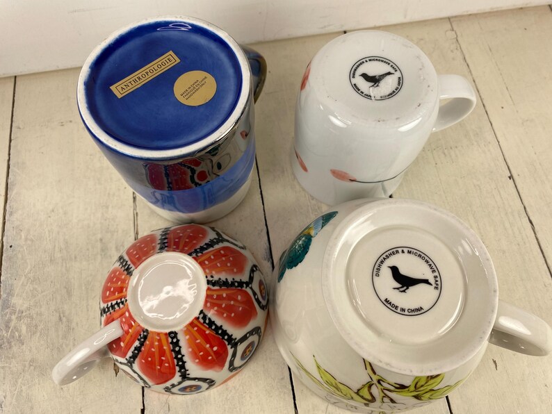 K&ouml;nnte beinhalten: Vier Keramikbecher mit verschiedenen Designs. Ein Becher hat ein blaues Innenleben mit dem Text "ANTHROPOLOGIE". Ein weiterer hat ein rot-oranges Blumenmuster. Die anderen beiden Becher sind wei&szlig; mit Vogel- und Blumendesigns. Alle Becher haben Griffe.