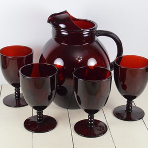 May include: Set of four ruby red glass wine glasses and a matching pitcher. The glasses have a stemmed base and a round bowl. The pitcher has a round body and a handle.