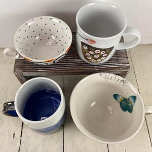 K&ouml;nnte beinhalten: Vier Keramikbecher und -schalen sind auf einer Holzoberfl&auml;che angeordnet. Eine Schale hat blaue Tupfen, eine andere ein Schmetterlingsdesign und eine dritte einen blauen Innenraum. Ein wei&szlig;er Becher zeigt ein Vogeldesign. Die Becher und Schalen sind wei&szlig; mit farbenfrohen Designs.