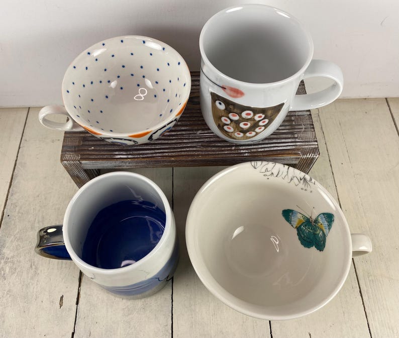K&ouml;nnte beinhalten: Vier Keramikbecher und -schalen sind auf einer Holzoberfl&auml;che angeordnet. Eine Schale hat blaue Tupfen, eine andere ein Schmetterlingsdesign und eine dritte einen blauen Innenraum. Ein wei&szlig;er Becher zeigt ein Vogeldesign. Die Becher und Schalen sind wei&szlig; mit farbenfrohen Designs.