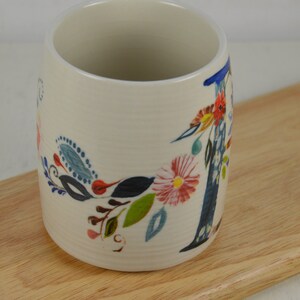 K&ouml;nnte beinhalten: Wei&szlig;e Keramiktasse mit einem Blumendesign mit blauen, gr&uuml;nen, rosa und gelben Blumen. Die Tasse hat einen gro&szlig;en Buchstaben "I" auf der Seite.