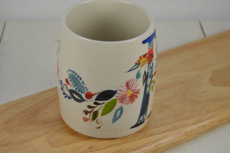 K&ouml;nnte beinhalten: Wei&szlig;e Keramiktasse mit einem Blumendesign mit blauen, gr&uuml;nen, rosa und gelben Blumen. Die Tasse hat einen gro&szlig;en Buchstaben "I" auf der Seite.