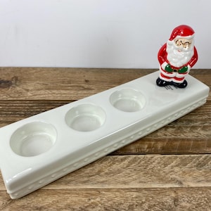 May include: A white ceramic candle holder with three circular indentations, and a decorative Santa Claus figurine. The candle holder has a rectangular shape with a beaded border. The Santa figurine is red, white, and black.