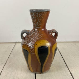 May include: A small, ceramic vase with a brown, yellow, and black design. The vase has two small handles and a gray rim. The body of the vase is a mix of colors, with a speckled texture on the upper portion.