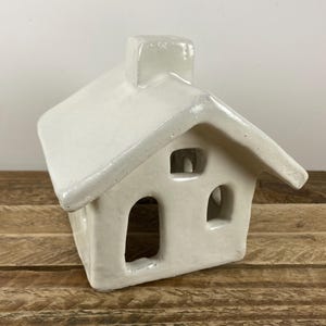 May include: A white ceramic house-shaped candle holder with a chimney and arched windows. The roof has a slight overhang, and the house sits on a wooden surface. The candle holder is a decorative item.