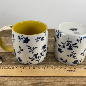 May include: Two ceramic mugs with a navy blue floral pattern on a white background. The inside of the mugs is yellow, and the handles are yellow with a geometric design. A small corgi figurine is next to the mugs.