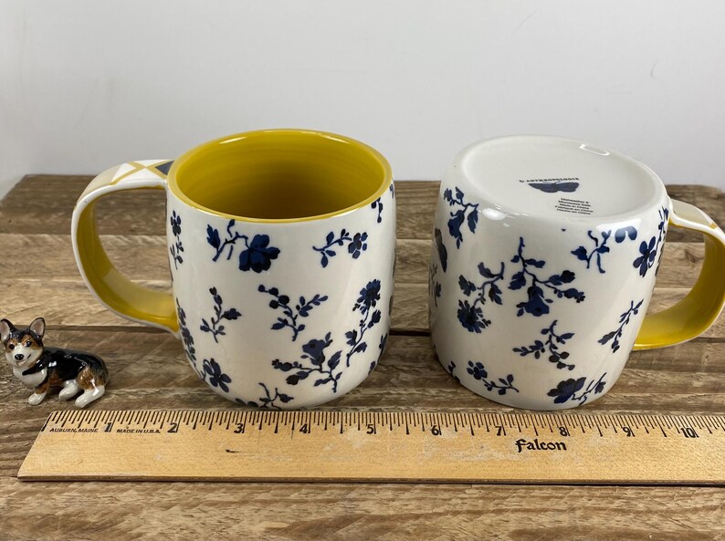 May include: Two ceramic mugs with a navy blue floral pattern on a white background. The inside of the mugs is yellow, and the handles are yellow with a geometric design. A small corgi figurine is next to the mugs.