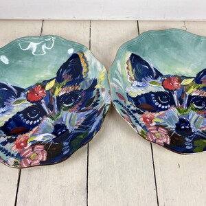 May include: Two decorative plates with a raccoon design. The plates feature a teal background with a raccoon face in the center, surrounded by colorful floral accents. The raccoon's fur is depicted in shades of blue, black, and white, with red and pink flowers.