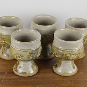May include: Set of five ceramic goblets with a white glaze and brown and green accents. The goblets have a footed base and a rounded bowl.