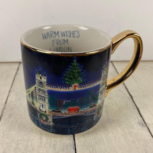 K&ouml;nnte beinhalten: Eine Keramik-Tasse mit goldenem Henkel und Rand. Die Tasse zeigt eine Nachtszene der Tower Bridge in London, mit einem Weihnachtsbaum und dem Text "Warm Wishes From London". Die Tasse ist dunkelblau.