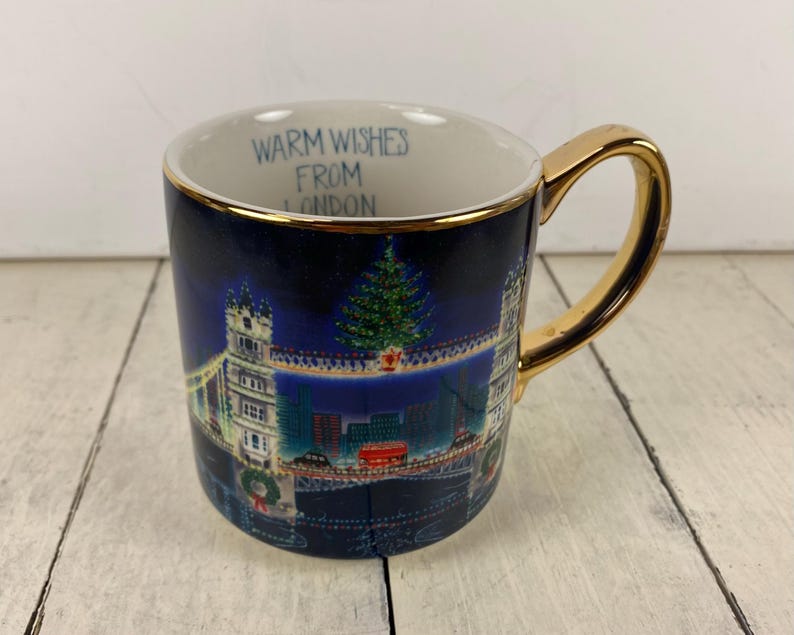 K&ouml;nnte beinhalten: Eine Keramik-Tasse mit goldenem Henkel und Rand. Die Tasse zeigt eine Nachtszene der Tower Bridge in London, mit einem Weihnachtsbaum und dem Text "Warm Wishes From London". Die Tasse ist dunkelblau.