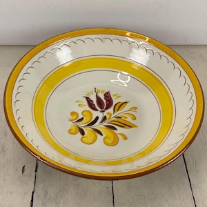 May include: A round, ceramic serving bowl with a white interior and a yellow and brown floral design. The bowl has a yellow rim and a scalloped edge. The exterior rim is brown.