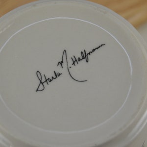 K&ouml;nnte beinhalten: Wei&szlig;e Keramiktasse mit einer schwarzen handschriftlichen Signatur, die am Boden "Carla M. Halfman" lautet.