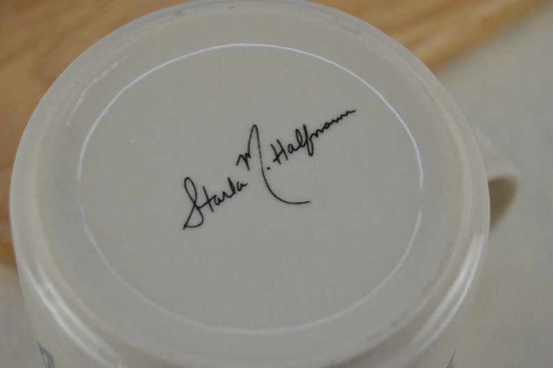 K&ouml;nnte beinhalten: Wei&szlig;e Keramiktasse mit einer schwarzen handschriftlichen Signatur, die am Boden "Carla M. Halfman" lautet.
