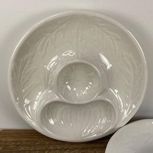 May include: A white ceramic serving dish with three compartments. The dish has a round shape with a central circular well and two crescent-shaped sections. The surface features a leaf-like embossed pattern. Another dish is partially visible.