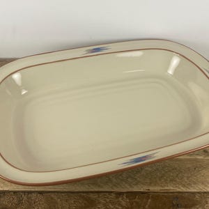 May include: Cream-colored rectangular serving dish with a brown rim and decorative blue and red geometric patterns. The dish has rounded corners and is set on a wooden surface.