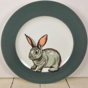 May include: A round ceramic plate with a teal rim and a white center. The center features a detailed illustration of a gray rabbit with pink inner ears. The rabbit is facing left.