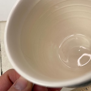 K&ouml;nnte beinhalten: Eine wei&szlig;e Keramiktasse mit cremefarbenem Innenraum. Die Tasse hat eine abgerundete Form und einen Henkel. Das Innere der Tasse hat konzentrische Ringe. Die Tasse wird von einer Hand gehalten.