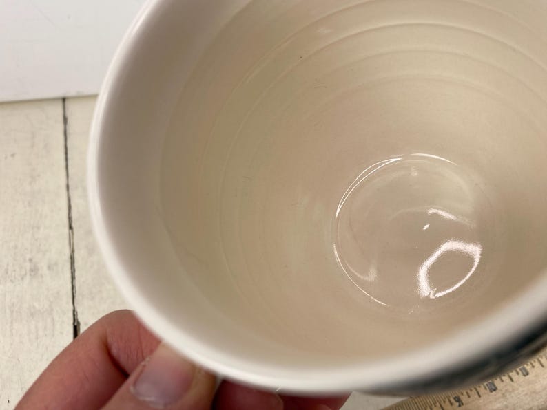 K&ouml;nnte beinhalten: Eine wei&szlig;e Keramiktasse mit cremefarbenem Innenraum. Die Tasse hat eine abgerundete Form und einen Henkel. Das Innere der Tasse hat konzentrische Ringe. Die Tasse wird von einer Hand gehalten.